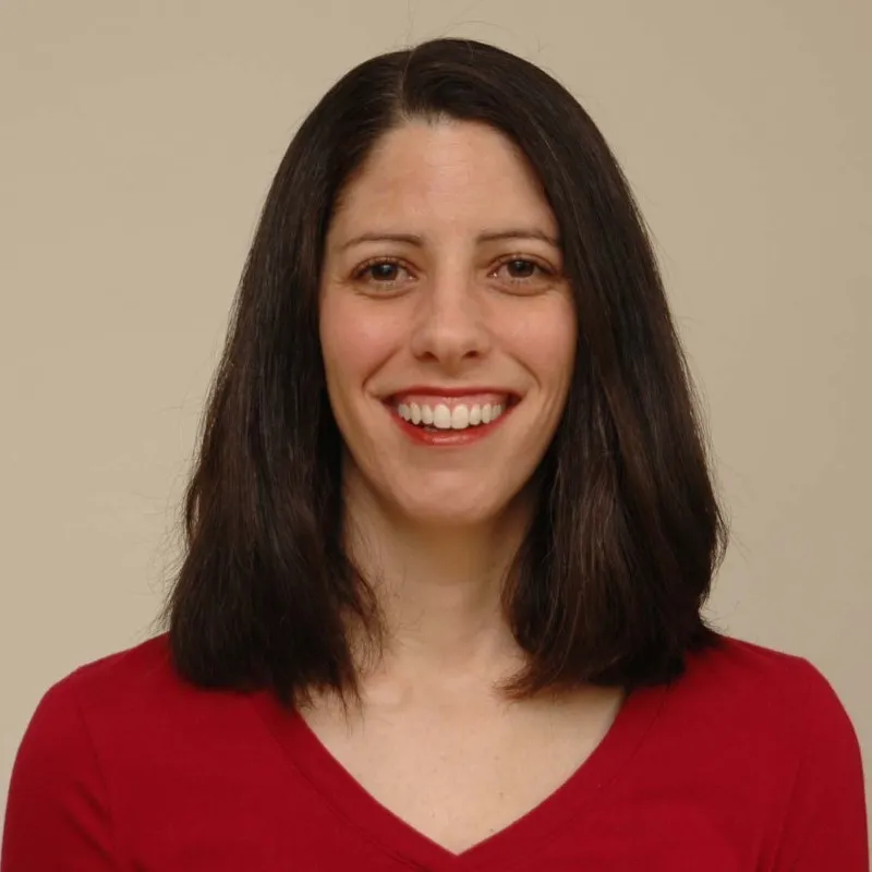 Quarter headshot of Amy, wearing a red v-neck shirt and a similar shade of lipstick, long brown hair parted slightly off to one side, and smiling towards the camera.