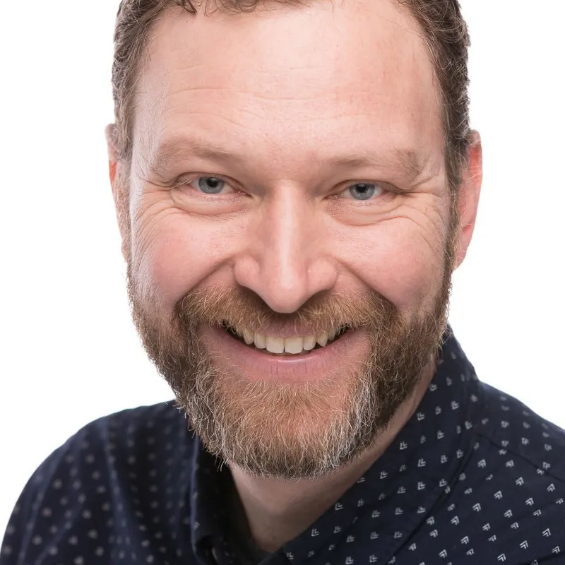 Quarter headshot of Ben, wearing a navy blue collared polka dotted shirt, brown hair and full beard, and smiling towards the camera.