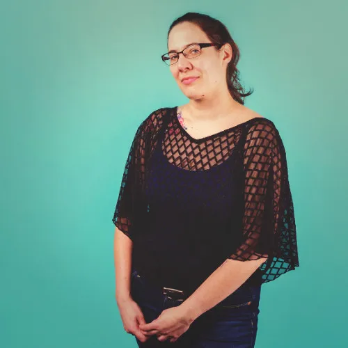 Half body shot of Catrina wearing blue jeans, a black mesh tee over a navy blue tank top, black rimmed glasses, and long dark brown hair pulled back. Catrina is positioned slightly to the right in front of a solid teal background, smiling directly at the camera with head tilted and hands lightly clasped in front.