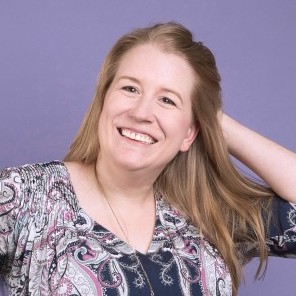 Quarter Headshot of Ericka in front of a light purple backdrop, wearing an intricately printed top and gold necklace, and smiling towards the camera with her hand in her long blondish brown hair behind her head.