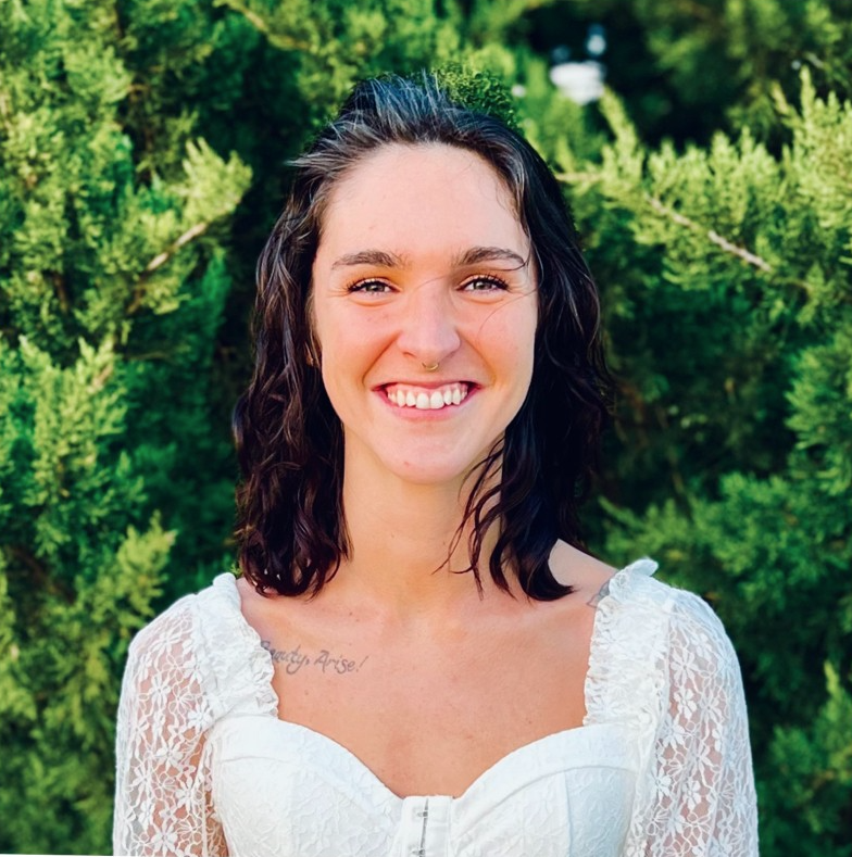 Quarter headshot of Michaela standing in front of some green evergreen trees, wearing a white dress, shoulder length hair, and smiling at the camera.
