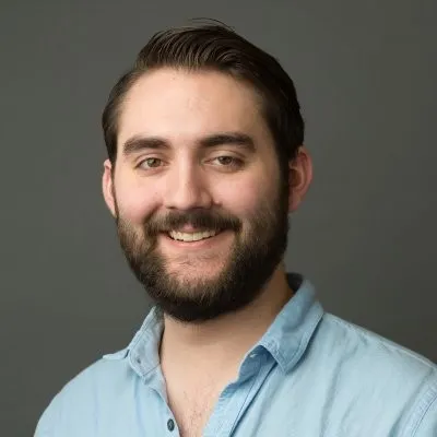 Qaurter headshot of Miguel in front of a grey backdrop, wearing a light blue button up collared shirt, brown hair parted on the side and combed back and a full brown beard, smiling towards the camera.