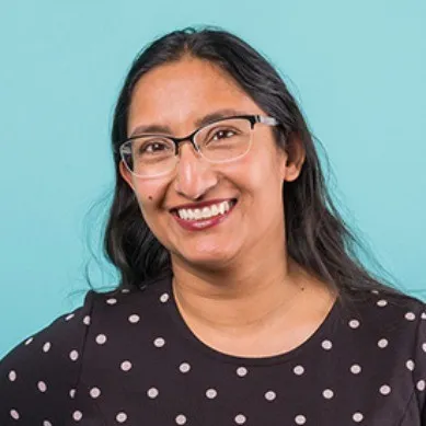 Quarter headshot of Sadia, wearing a dark shirt with light polka dots and horn rimmed glasses, long black hair parted down the middle, and smiling towards the camera.